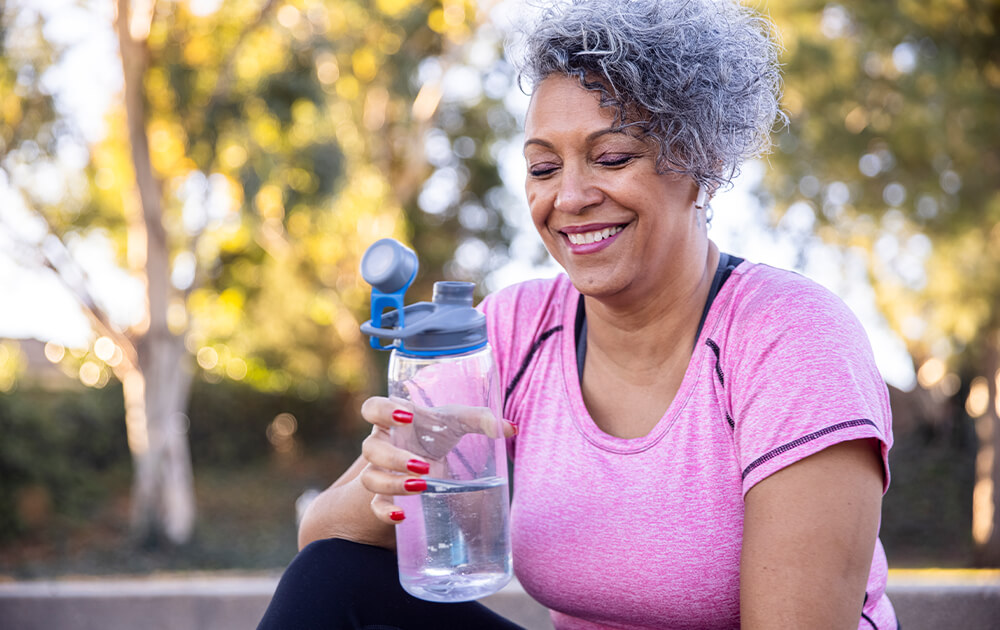 A happy, older woman drinks from a water bottle outside after a workout.