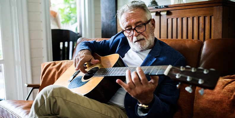 Elderly man in glasses playing an acoustic guitar on a brown leather couch.