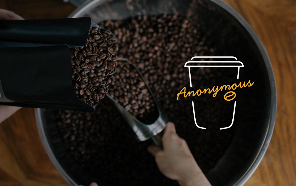 Coffee beans being poured into a large container with a coffee cup outline and 'Anonymous' written overlay. 