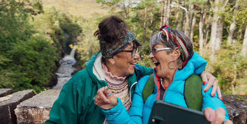 Two older women taking a selfie outdoors with a forest and waterfall in the background.