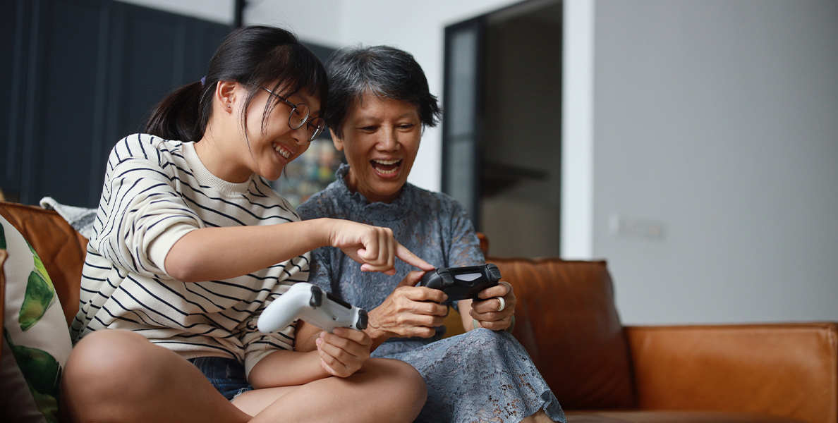 An older woman and her adult daughter sit on a couch playing video games together, both smiling and holding game controllers.
