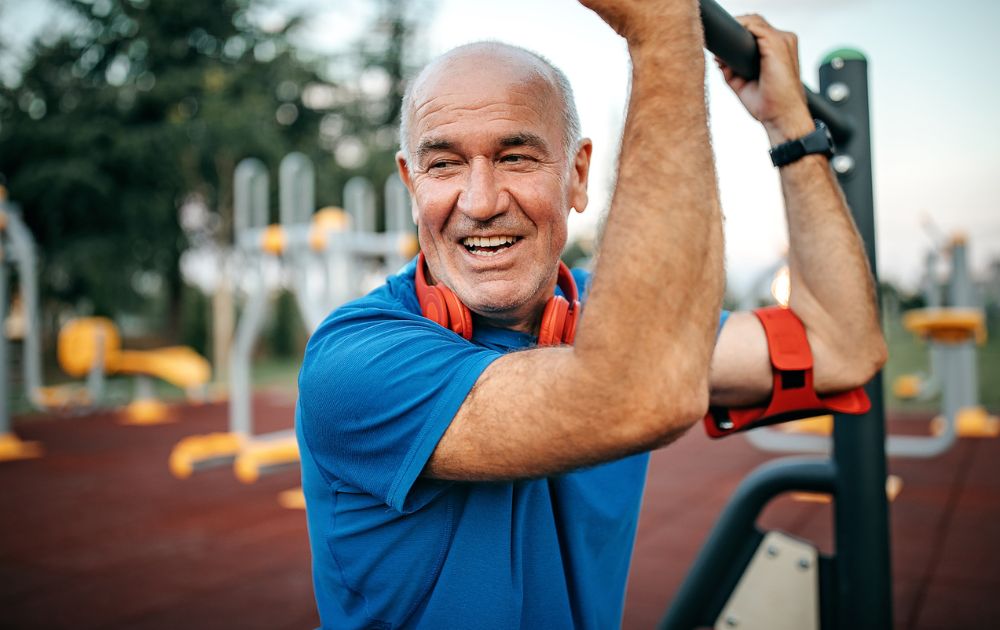 An older man smiling while exercising on outdoor gym equipment.