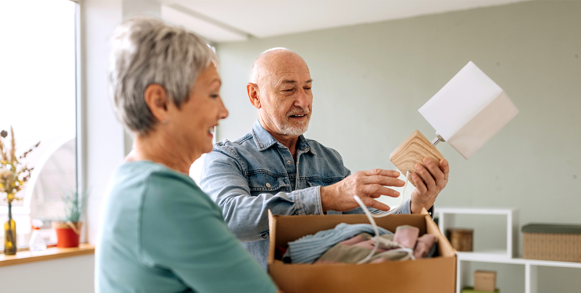  smiling senior couple unpacking a moving box together in a new home; the man holds up a small wooden lamp.