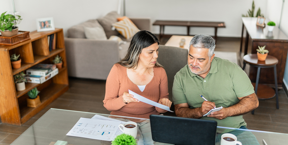 A middle-aged couple sitting at their dining table with a laptop and papers, seriously discussing their finances.