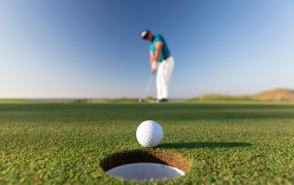 A golf ball rests on the edge of the cup on a putting green, with a golfer in the background.