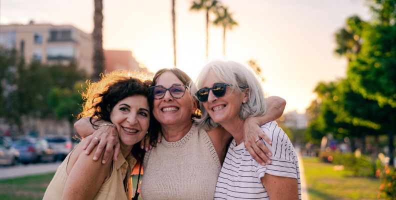 Three smiling senior women with their arms around each other, standing outdoors at sunset