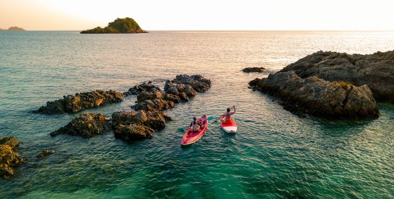 Two people paddleboard and kayak through calm, turquoise waters near rocky outcrops at sunset.