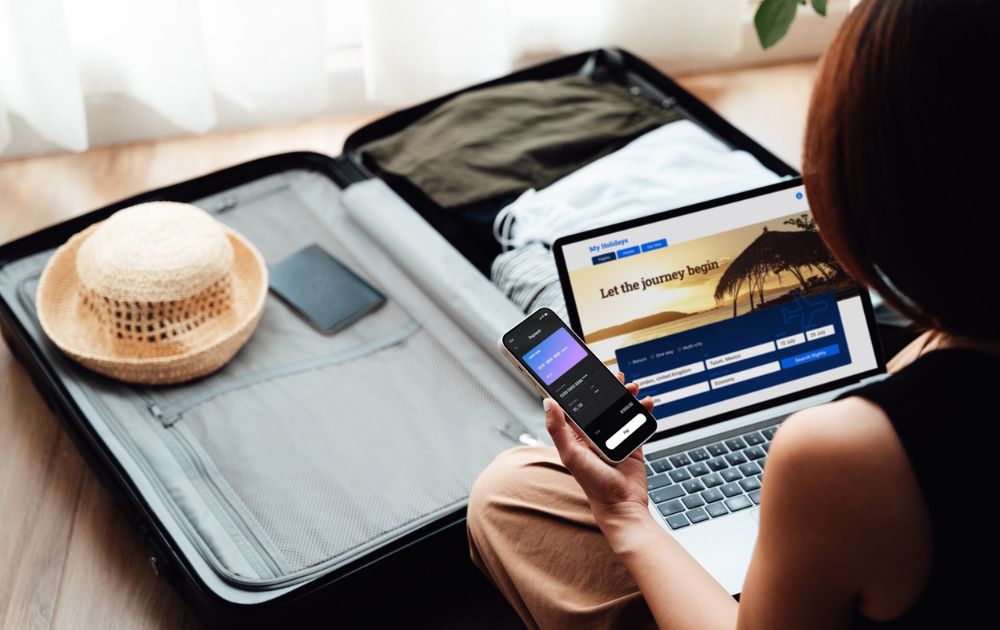 A person sits on the floor next to an open suitcase while using a laptop and smartphone to book a trip.