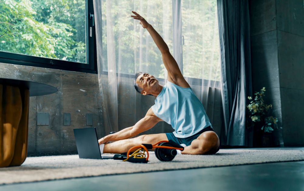 An older man performs a seated side stretch on a yoga mat in a bright, modern room while following a video on his laptop.