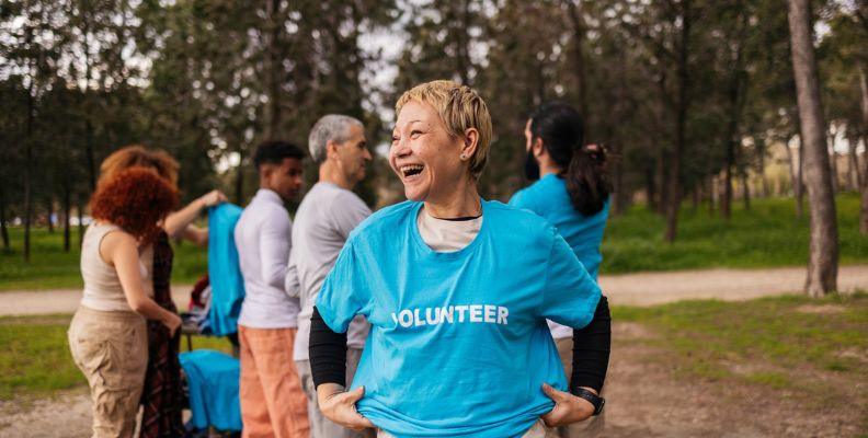 A woman in a blue volunteer t-shirt laughs joyfully while standing with a group of people outdoors.