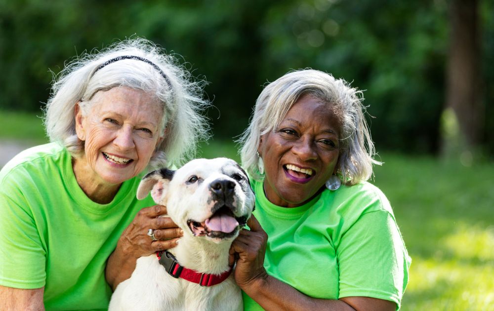 Two smiling women in matching lime green shirts pose closely with a happy white dog in a park.