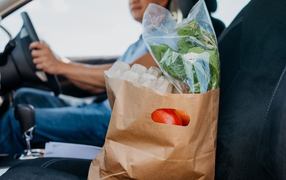 A brown paper grocery bag filled with eggs and fresh greens sits on the passenger seat of a car while a person drives.