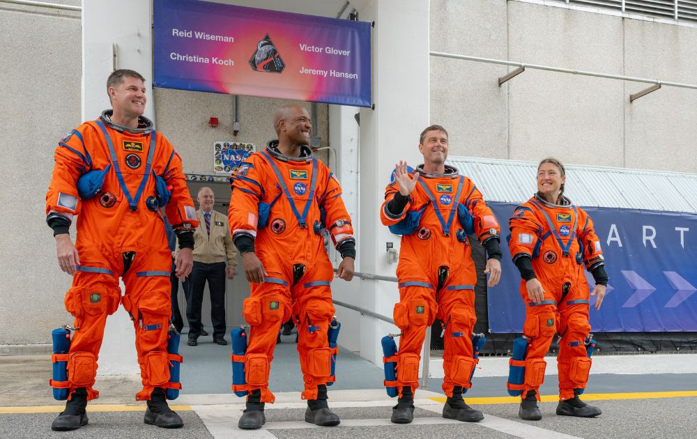 The four Artemis II astronauts in bright orange spacesuits wave and smile while walking outside the Kennedy Space Center.