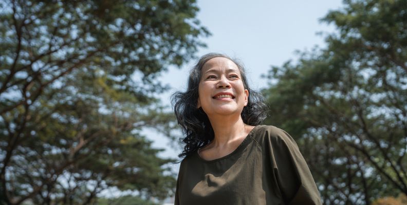 A smiling woman looks upward with a positive expression against a backdrop of leafy green trees and a clear sky.