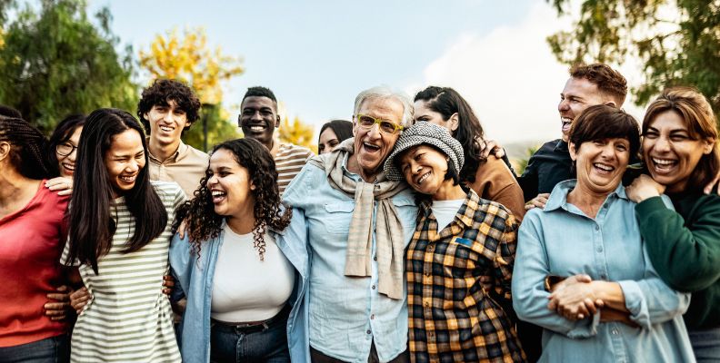 A large, diverse group of happy friends and family members of all ages laughing and hugging outdoors.