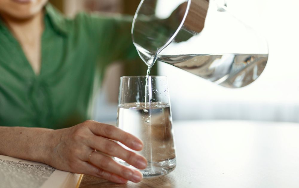 A close-up of a person pouring fresh, clear water from a glass pitcher into a drinking glass on a wooden table.