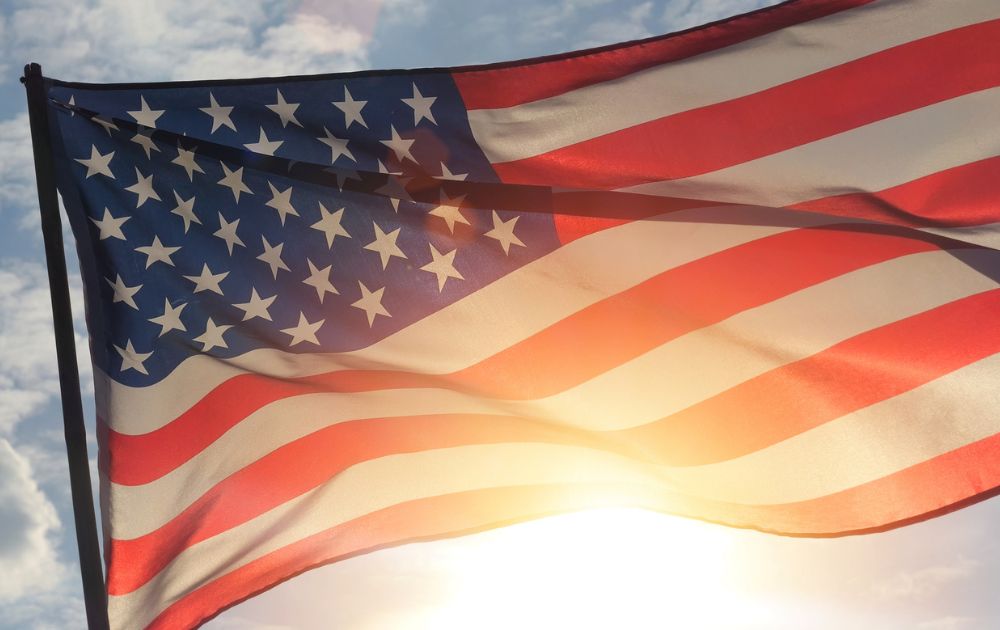 An American flag waves in the breeze against a bright blue sky with sunlight glowing through the fabric.