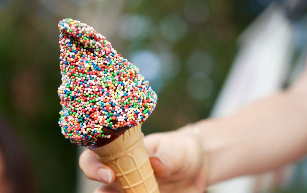 A child's hand holds a waffle cone covered in colorful rainbow sprinkles against a blurred green background.