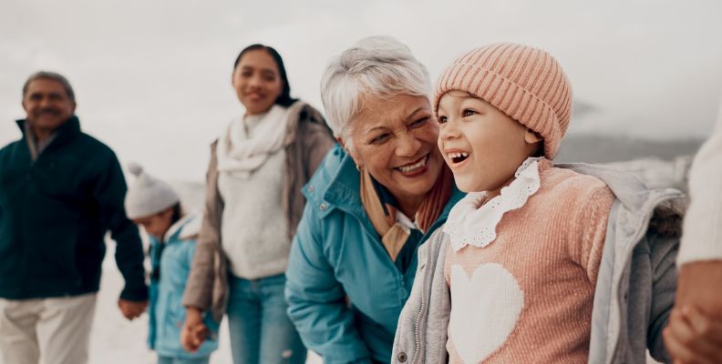 A smiling grandmother leans in close to a laughing young child while family members walk together on a beach in cool weather.