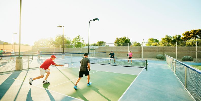 Four adults play doubles pickleball on an outdoor court in bright morning sunlight.